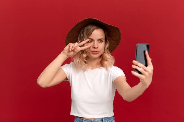 Beyaz bir t-shirt ve günbatımı şapka kıvırcık saçlı sarışın kız kırmızı bir arka plan üzerinde cep telefonu ile selfie yapar.