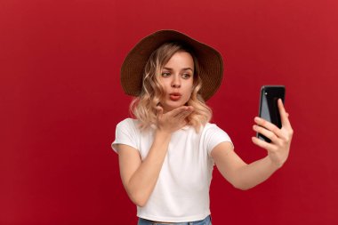 Beyaz bir t-shirt ve günbatımı şapka kıvırcık saçlı sarışın kız kırmızı bir arka plan üzerinde cep telefonu ile selfie yapar.
