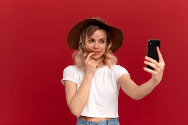Beyaz bir t-shirt ve günbatımı şapka kıvırcık saçlı sarışın kız kırmızı bir arka plan üzerinde cep telefonu ile selfie yapar.