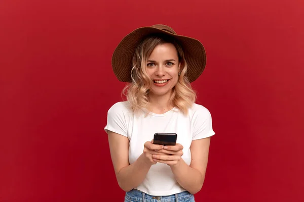 白いTシャツに巻き毛のブロンドの髪をした少女の肖像画と、赤い背景に日没の帽子。幸せなモデルは、携帯電話の画面を見て. — ストック写真