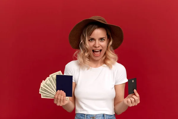 Lycklig tjej med lockigt blont hår i en vit t-shirt och en solhatt på en röd bakgrund. Modell innehar pass med kontanter och ett bankkort. — Stockfoto