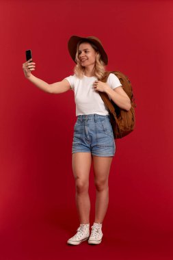 Kıvırcık sarı saçlı bir kızın portresi, güneşli şapka ve beyaz tişört giymiş kırmızı bir arka plan üzerinde durarak cep telefonuyla selfie çekerken. Model turuncu sırt çantası ile seyahat.