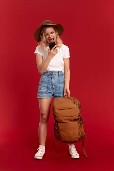 Troubled girl with curly blond hair dressed in a sundwon hat and white t-shirt standing on a red background video chatting with mobile phone. Model travels with orange backpack. — Stock Photo, Image