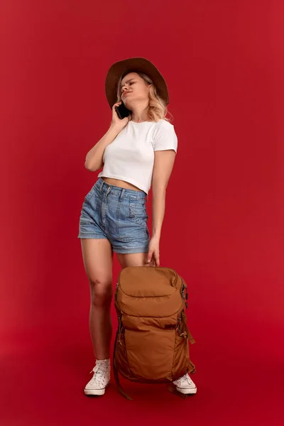 Troubled girl with curly blond hair dressed in a sundwon hat and white t-shirt standing on a red background video chatting with mobile phone. Model travels with orange backpack. — Stock Photo, Image