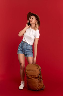 Kıvırcık sarı saçlı sorunlu kız bir sundwon şapka ve beyaz t-shirt kırmızı bir arka plan video cep telefonu ile sohbet ayakta giymiş. Model turuncu sırt çantası ile seyahat.