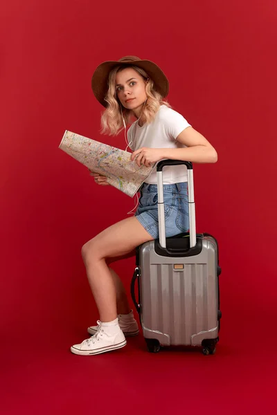 Młoda blond kręcone kobieta siedzi na szarej torbie bagażowej z mapą przed czerwonym tłem. — Zdjęcie stockowe