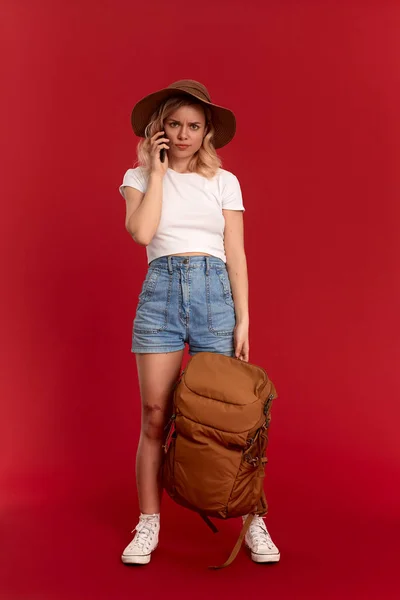 Troubled girl with curly blond hair dressed in a sundwon hat and white t-shirt standing on a red background video chatting with mobile phone. Model travels with orange backpack. — Stock Photo, Image