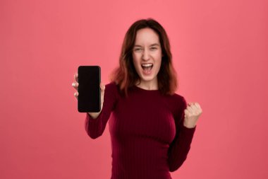 Heyecanlı güzel esmer kız kameraya bakarak pembe bir arka plan üzerinde dururken bir cep telefonu ekran gösteren.