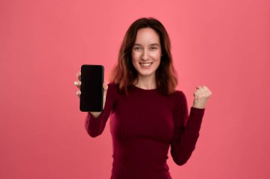 Heyecanlı güzel esmer kız kameraya bakarak pembe bir arka plan üzerinde dururken bir cep telefonu ekran gösteren.