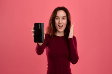 Heyecanlı güzel esmer kız kameraya bakarak pembe bir arka plan üzerinde dururken bir cep telefonu ekran gösteren.