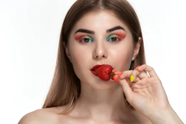 Retrato de close-up de uma bela jovem com maquiagem colorida brilhante segurando morango na boca . — Fotografia de Stock
