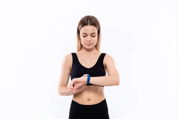 Joven mujer rubia deportiva en una ropa deportiva negra mirando los relojes inteligentes comprobando el pulso después del entrenamiento de pie sobre fondo blanco . — Foto de Stock