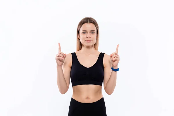 Ung sportig blond kvinna i en svart sportkläder med smarta klockor för pulsmätning pekar med fingrarna upp. — Stockfoto