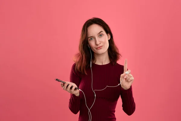 Retrato de uma menina bonita morena segurando fone de ouvido em uma mão e telefone celular em outra em pé sobre fundo rosa . — Fotografia de Stock