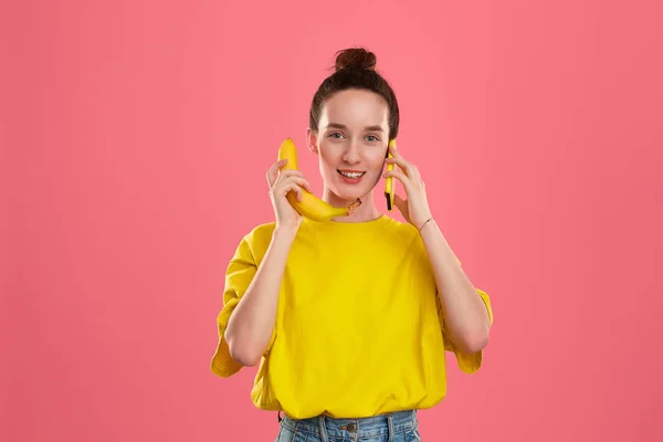 Modelo sonriente con un moño de pelo en una camiseta amarilla que sostiene plátano y teléfono inteligente amarillo — Foto de Stock
