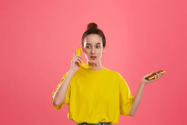 Modelo confuso con un moño de pelo en una camiseta amarilla sosteniendo plátano hablando en un teléfono inteligente amarillo — Foto de Stock