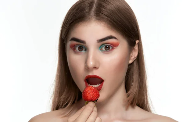 Retrato de close-up de uma bela jovem com maquiagem colorida brilhante segurando morango perto do rosto . — Fotografia de Stock