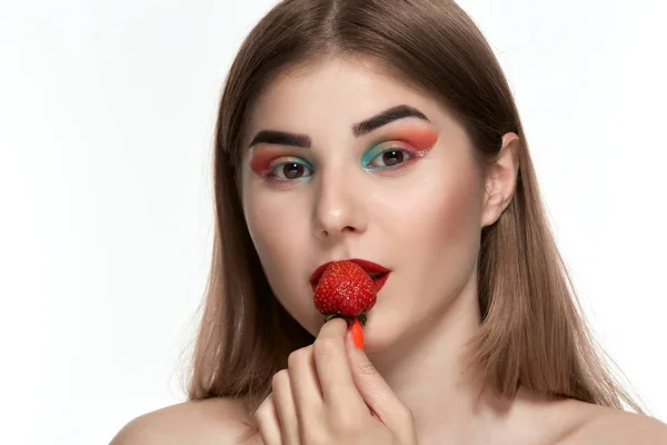 Retrato de cerca de una hermosa joven con maquillaje de color brillante sosteniendo fresa cerca de la cara . — Foto de Stock