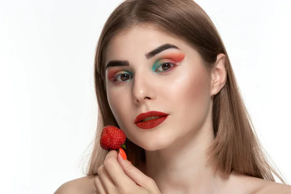 Retrato de cerca de una hermosa joven con maquillaje de color brillante sosteniendo fresa cerca de la cara . — Foto de Stock