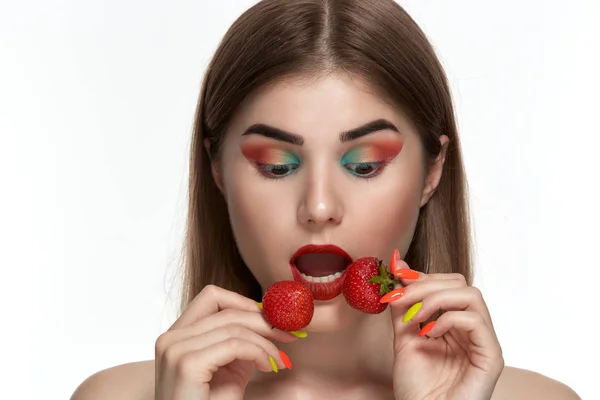 Retrato de close-up de uma bela jovem com maquiagem colorida brilhante segurando morango perto do rosto . — Fotografia de Stock