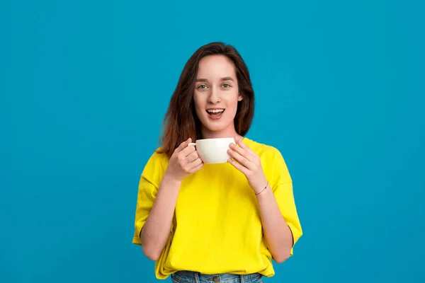 Hübsche brünette Frau in einem gelben T-Shirt, die Getränke aus einer weißen Tasse trinkt, die vor blauem Hintergrund steht. — Stockfoto