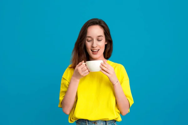 Mulher morena bonita em uma camiseta amarela bebendo bebida de uma xícara branca em pé sobre fundo azul . — Fotografia de Stock