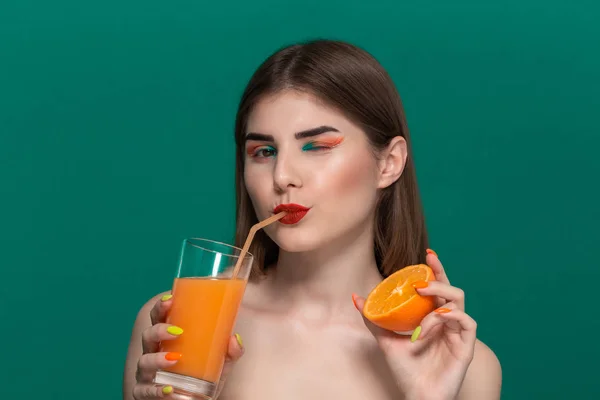 Retrato de close-up de bela jovem com maquiagem de cor brilhante bebendo suco de laranja e segurando metade de uma laranja — Fotografia de Stock
