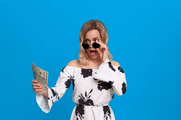 Retrato de una chica con el pelo rubio rizado en un vestido blanco y gafas de sol oscuras de pie sobre un fondo azul mirando sorprendentemente el paquete de dólares . —  Fotos de Stock