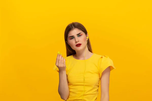Ung modell i en gul tee har problem med att lyssna på musik med kabelanslutna hörlurar stående över gul bakgrund. — Stockfoto