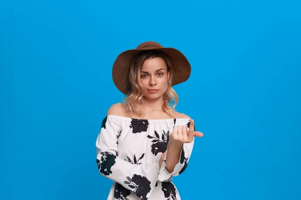 Gruñón joven modelo con pelo rubio rizado en un sombrero de puesta del sol y vestido blanco invita con un dedo de pie sobre un fondo azul . —  Fotos de Stock