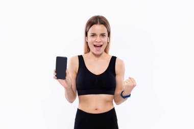 Beyaz arka plan üzerinde duran bir parmak ile bir akıllı telefon ekran gösteren kablosuz kulak tomurcukları ile siyah bir spor giyim heyecanlı genç sportif sarışın kadın.