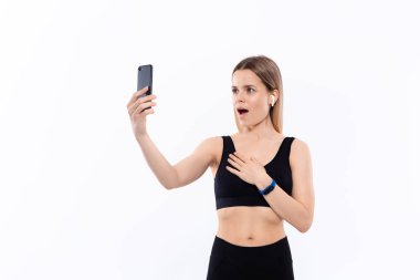 Siyah spor giyimli genç sportif sarışın kadın beyaz arka plan üzerinde duran egzersiz sonrası pazı gösteren akıllı telefon ile selfie yapıyor. Model kablosuz kulaklıkile egzersiz sırasında müzik dinler