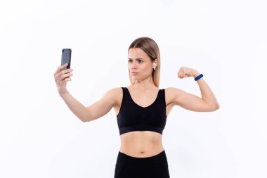 Siyah spor giyimli genç sportif sarışın kadın beyaz arka plan üzerinde duran egzersiz sonrası pazı gösteren akıllı telefon ile selfie yapıyor. Model kablosuz kulaklıkile egzersiz sırasında müzik dinler