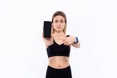 Kablosuz kulak tomurcukları beyaz arka plan üzerinde duran bir parmak ile bir akıllı telefon ekran gösteren siyah bir spor giyim genç sportif sarışın kadın.