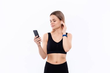 Kablosuz kulak tomurcukları ile siyah bir spor giyim heyecanlı genç sportif sarışın kadın beyaz arka plan üzerinde izole duran bir akıllı telefon ekranına bakıyor.