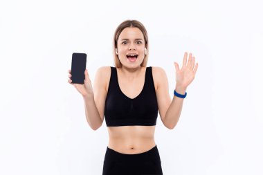Beyaz arka plan üzerinde duran bir parmak ile bir akıllı telefon ekran gösteren kablosuz kulak tomurcukları ile siyah bir spor giyim heyecanlı genç sportif sarışın kadın.
