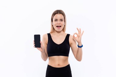 Beyaz arka plan üzerinde duran bir parmak ile bir akıllı telefon ekran gösteren kablosuz kulak tomurcukları ile siyah bir spor giyim heyecanlı genç sportif sarışın kadın.