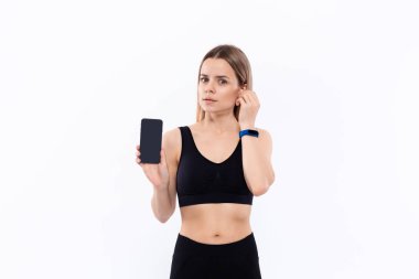 Kablosuz kulak tomurcukları beyaz arka plan üzerinde duran bir parmak ile bir akıllı telefon ekran gösteren siyah bir spor giyim genç sportif sarışın kadın.