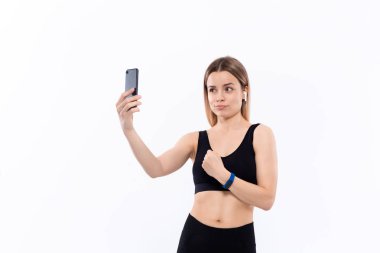 Siyah spor giyimli genç sportif sarışın kadın beyaz arka plan üzerinde duran egzersiz sonrası pazı gösteren akıllı telefon ile selfie yapıyor. Model kablosuz kulaklıkile egzersiz sırasında müzik dinler