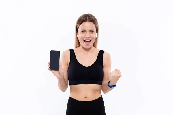 Upphetsad ung sportig blond kvinna i en svart sportkläder med trådlösa öronsnäckor visar skärmen på en smartphone med ett finger stående över vit bakgrund. — Stockfoto