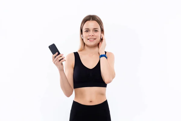 Mujer rubia deportiva joven excitada en una ropa deportiva negra con auriculares inalámbricos mirando la pantalla de un teléfono inteligente de pie aislado sobre fondo blanco . —  Fotos de Stock