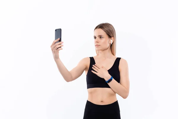 Jeune femme blonde sportive dans un selfie de vêtements de sport noirs avec smartphone montrant biceps après l'entraînement debout sur fond blanc. Modèle écoute de la musique pendant l'entraînement avec casque sans fil — Photo