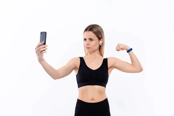 Ung sportig blond kvinna i en svart sportkläder göra Selfie med smartphone visar bicep efter träningen stående över vit bakgrund. Modell lyssnar musik under träningen med trådlösa hörlurar — Stockfoto