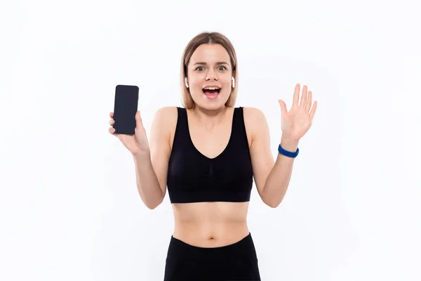 Podekscytowany młoda sportowa blond kobieta w czarnej odzieży sportowej z bezprzewodowych słuchawek pokazano ekran Smartphone z palcem stoi na białym tle. — Zdjęcie stockowe