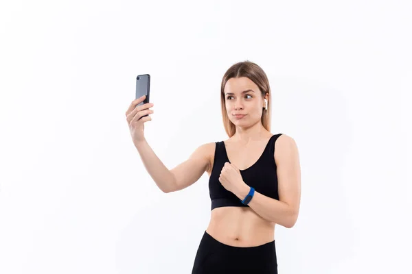 Jeune femme blonde sportive dans un selfie de vêtements de sport noirs avec smartphone montrant biceps après l'entraînement debout sur fond blanc. Modèle écoute de la musique pendant l'entraînement avec casque sans fil — Photo