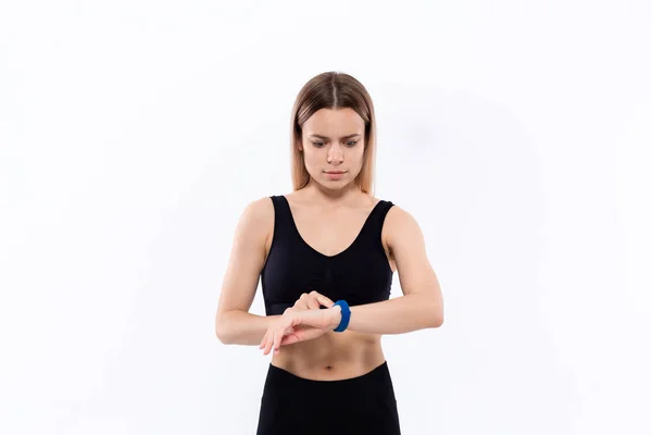 Joven mujer rubia deportiva en una ropa deportiva negro sorprendido comprobar el pulso con relojes inteligentes después del entrenamiento de pie sobre fondo blanco — Foto de Stock