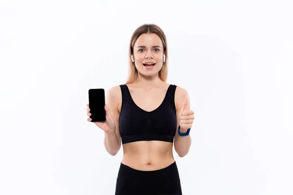 Upphetsad ung sportig blond kvinna i en svart sportkläder med trådlösa öronsnäckor visar skärmen på en smartphone med ett finger stående över vit bakgrund. — Stockfoto