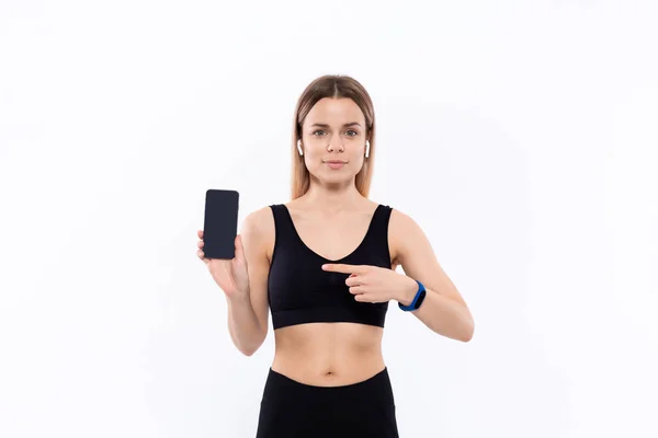 Ung sportig blond kvinna i en svart sportkläder med trådlösa öronsnäckor visar skärmen på en smartphone med ett finger stående över vit bakgrund. — Stockfoto