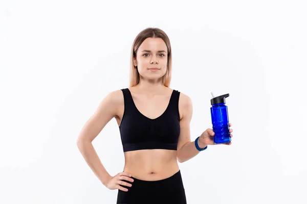 Fiatal sportos szőke nő egy fekete sportruházat intelligens órák tartja palack vízzel edzés után álló fehér háttér. — Stock Fotó