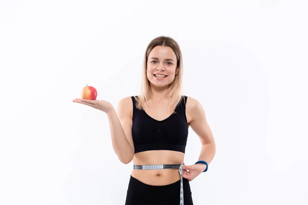 Ung blond kvinna i en svart sportkläder med smarta klockor är glad att kontrollera midjan efter träningen med måttband som innehar rött äpple stående över vit bakgrund. — Stockfoto